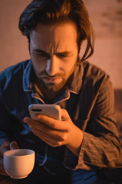 Чоловік з розладом сну використовує смартфон і тримає чашку кави в спальні — стокове фото