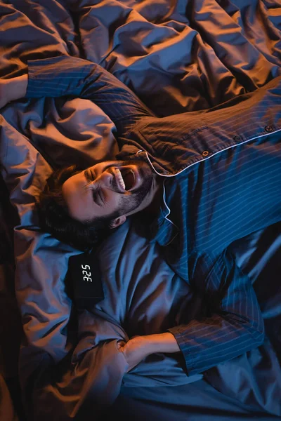 Vista superior do homem com insônia bocejando enquanto deitado na cama perto do despertador à noite — Fotografia de Stock