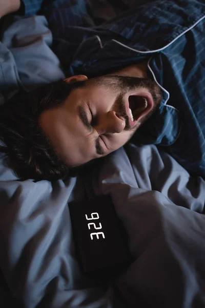 Top view of sleepless man yawning near alarm clock on bed at night — Stock Photo