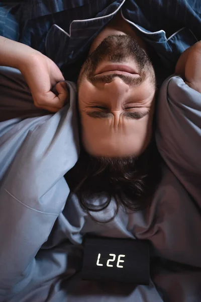 Top view of tensed man covering ears near alarm clock on bed at night — Photo de stock