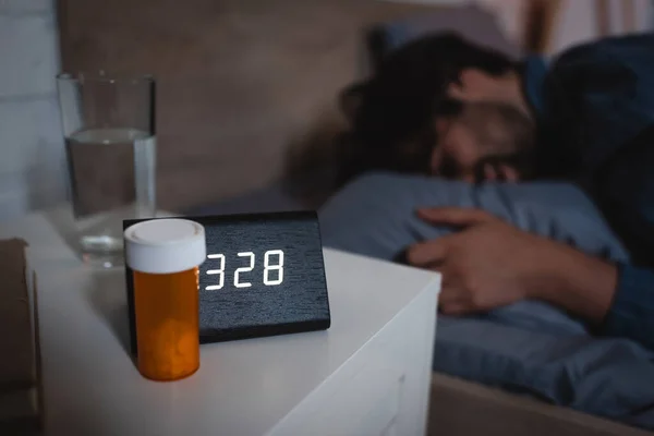 Pills and alarm clock on bedside table near blurred man on bed at night — Stock Photo