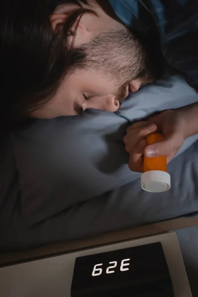 Vista lateral del joven con trastorno del sueño sosteniendo pastillas cerca del reloj despertador en el dormitorio por la noche - foto de stock