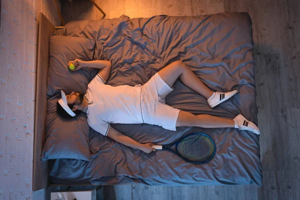 Side view of man in sportswear holding tennis ball and rocket while sleeping at home — Fotografia de Stock