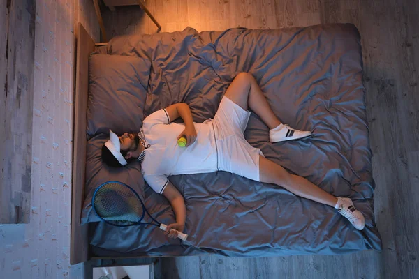 Joven sosteniendo cohete de tenis y pelota mientras duerme en la cama en casa - foto de stock