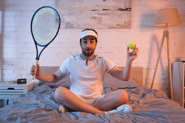 Amazed somnambulist in sportswear holding tennis ball and rocket on bed at night — Fotografia de Stock