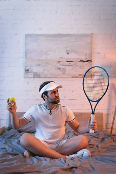 Confused sleepwalker in sportswear holding tennis ball and rocket on bed — Stockfoto