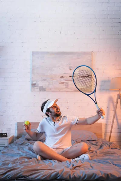 Emocionado sonámbulo en ropa deportiva sosteniendo cohete de tenis y pelota en la cama por la noche - foto de stock