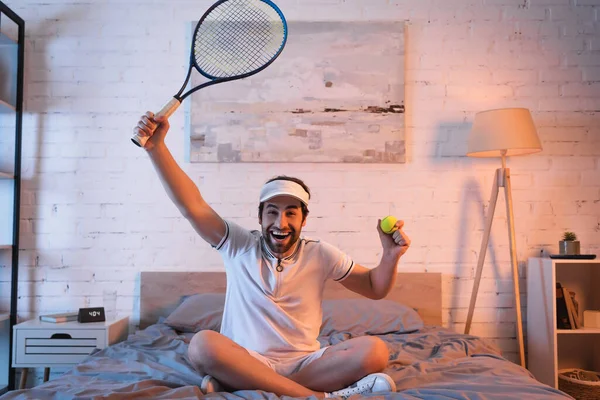 Cheerful sleepwalker in sportswear holding tennis rocket and ball on bed — Stock Photo