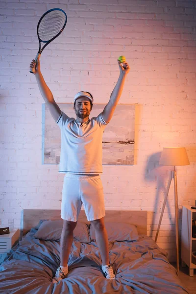 Emocionado sonámbulo sosteniendo pelota de tenis y cohete en la cama por la noche - foto de stock