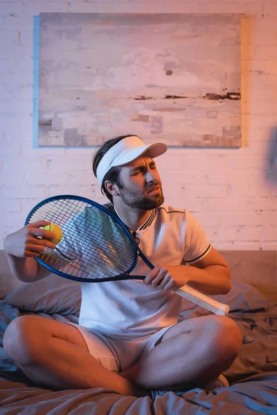 Confused sleepwalker holding tennis racket and ball while sitting on bed at night — Fotografia de Stock