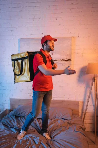 Smiling deliveryman outstretching hand while suffering from sleepwalking on bed - foto de stock