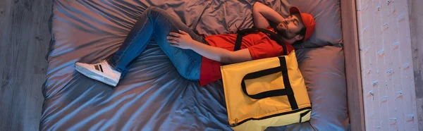 Top view of young somnambulist in delivery uniform sleeping near thermo backpack on bed, banner - foto de stock