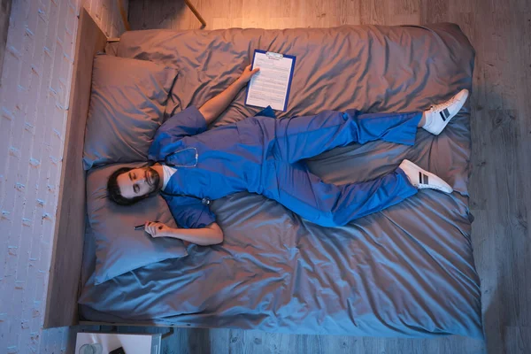 Top view of sleepwalker in doctor uniform holding clipboard and pen while sleeping on bed — Stock Photo
