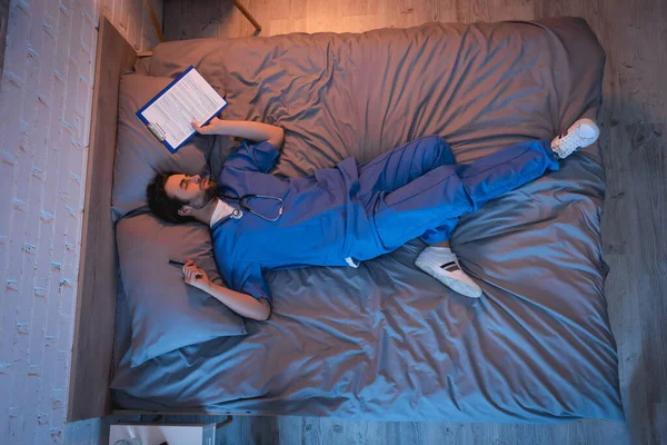 Молодой лунатик в форме доктора держит ручку и планшет во время сна на кровати — стоковое фото