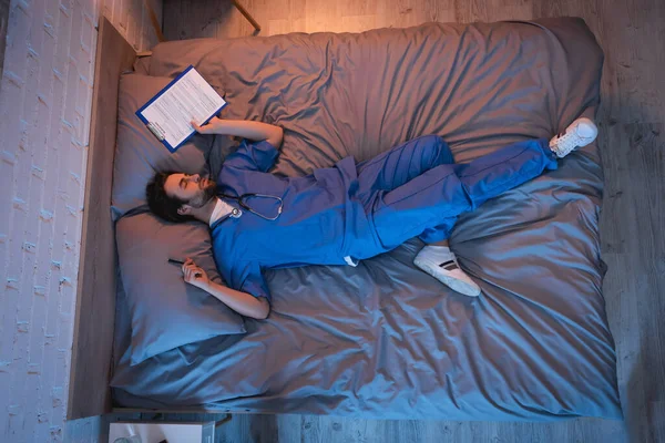 Young sleepwalker in doctor uniform holding pen and clipboard while sleeping on bed — Stock Photo