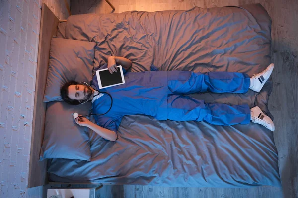Top view of sleepwalker in doctor uniform holding digital tablet and stethoscope while sleeping on bed — Stock Photo
