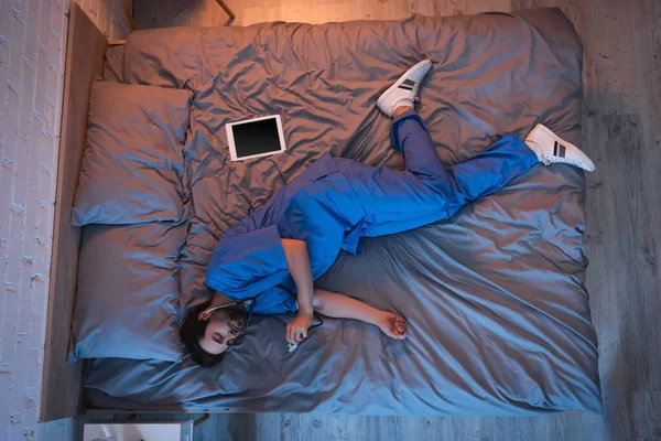 Вид спящего человека в форме врача, держащего стетоскоп рядом с цифровым столиком на кровати дома — стоковое фото