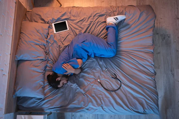 Вид сбоку на молодого лунатика в форме врача, спящего возле цифрового планшета и стетоскопа на кровати — стоковое фото