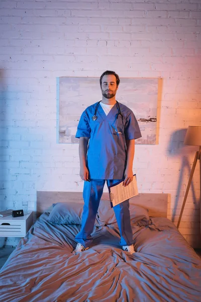 Sleepwalker in doctor uniform holding clipboard while standing on bed at home — Stock Photo