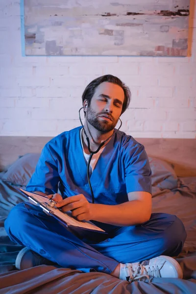 Sleepwalker in doctor uniform and stethoscope pointing at clipboard on bed at night — Stock Photo