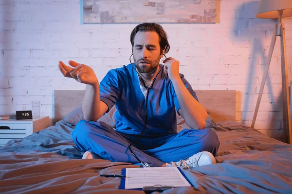 Sleepwalker in doctor uniform and stethoscope gesturing near clipboard on bed at night — Stock Photo