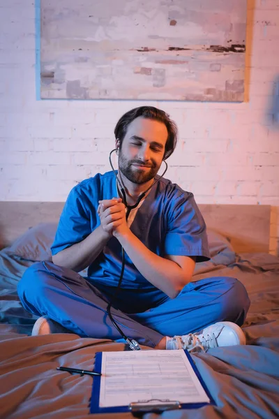 Smiling somnambulist in doctor uniform and stethoscope sitting near clipboard on bed — Stock Photo