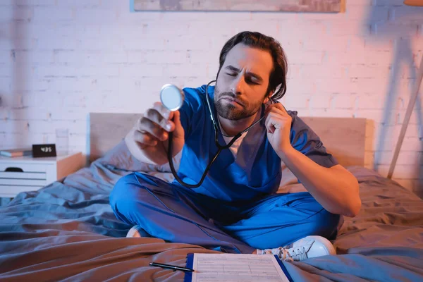 Schlafwandler in Arztuniform hält nachts Stethoskop in der Nähe des Klemmbretts auf dem Bett — Stockfoto