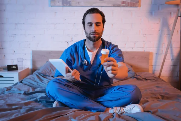 Sleepwalker in doctor uniform holding pills and digital tablet on bed - foto de stock