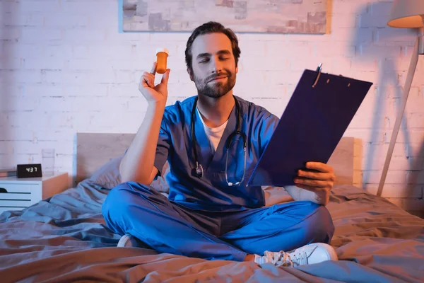 Smiling sleepwalker in doctor uniform holding clipboard and pills on bed at night — Stock Photo