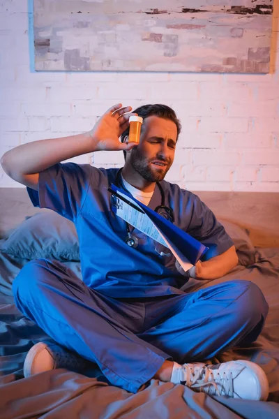Worried sleepwalker in doctor uniform holding clipboard and pills on bed at night — Photo de stock