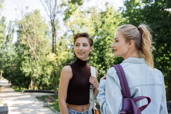 Cheerful students holding backpacks and looking away in park — Stock Photo