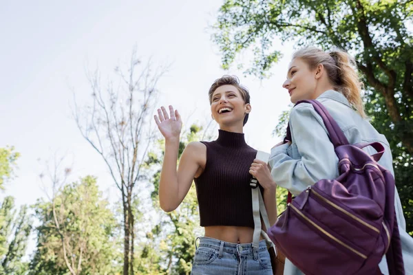 Low angle view of student waving hand near friend in park — Stock Photo