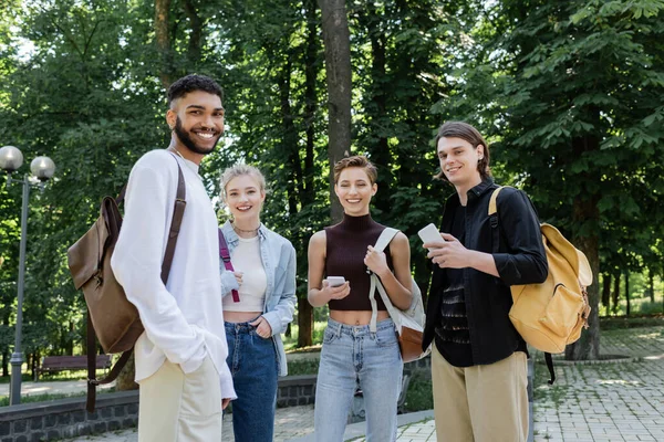 Studenti interrazziali sorridenti con zaini e smartphone che guardano la fotocamera nel parco — Foto stock