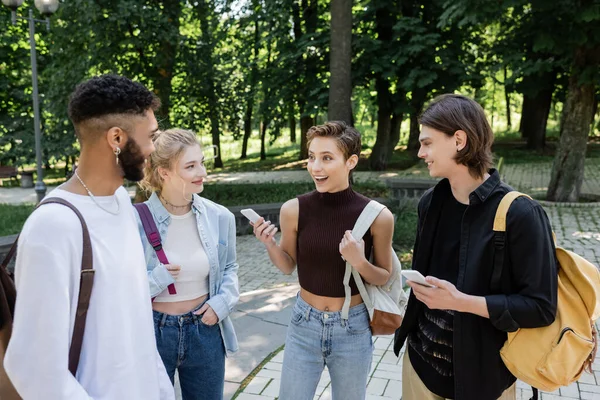 Веселий студент зі смартфоном розмовляє з міжрасовими друзями в парку — стокове фото