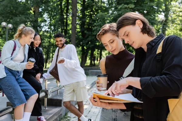 Estudiantes mirando cuaderno cerca borrosa amigos multiétnicos con café y teléfonos inteligentes en el parque - foto de stock