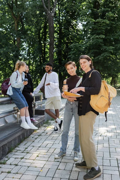 Des étudiants heureux tenant des cahiers près d'amis multiculturels flous dans le parc — Photo de stock