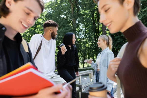 Lächelnde multiethnische Freunde bei Kaffee und Smartphone, die sich in der Nähe verschwommener Freunde im Park unterhalten — Stockfoto