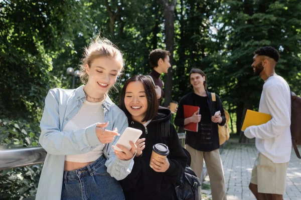 Улыбающийся студент указывает пальцем на смартфон рядом с азиатским другом с кофе, чтобы пойти в парк — стоковое фото