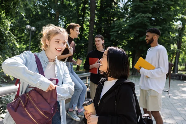 Positive interracial students with coffee to go talking near blurred friends in park — Stock Photo