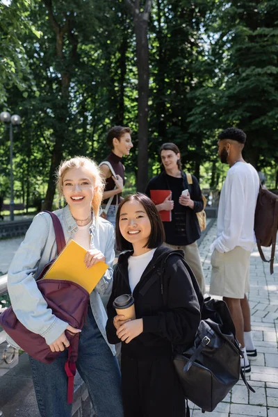 Étudiants interraciaux avec cahier et café pour aller sourire à la caméra près des amis flous dans le parc — Photo de stock