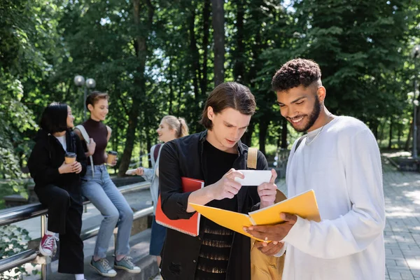 Sonriente estudiante afroamericano sosteniendo un cuaderno mientras un amigo toma una foto en un teléfono inteligente en el parque - foto de stock