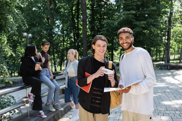 Estudiantes multiétnicos positivos con cuadernos y teléfonos inteligentes mirando a la cámara en el parque - foto de stock