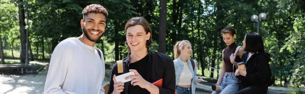 Cheerful interracial students with notebook and smartphone standing near friends in park, banner - foto de stock