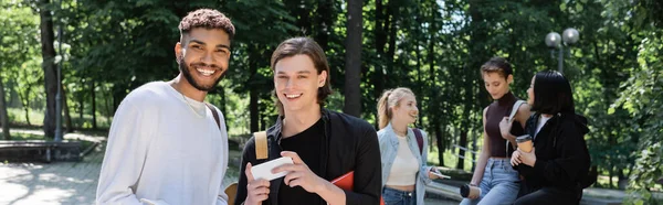 Étudiants interracial joyeux avec ordinateur portable et smartphone debout près des amis dans le parc, bannière — Photo de stock