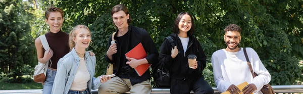 Cheerful interracial students with backpacks and notebooks looking at camera in park, banner - foto de stock