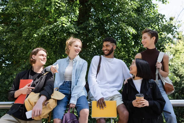 Positive multicultural students with notebooks talking in park — Foto stock