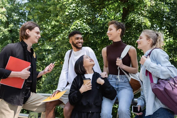 Улыбающиеся мультикультурные студенты с ноутбуками и смартфонами разговаривают в парке — стоковое фото