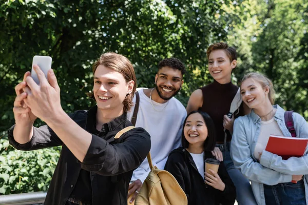 Estudiante sonriente tomando selfie en smartphone cerca de amigos interracial en el parque - foto de stock