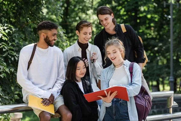 Fröhliche multiethnische Studenten schauen im Park auf Notizbuch — Stockfoto