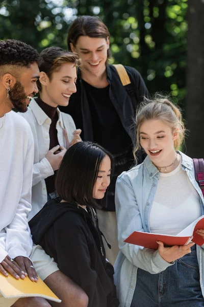 Estudiante sosteniendo cuaderno cerca de alegres amigos multiculturales - foto de stock